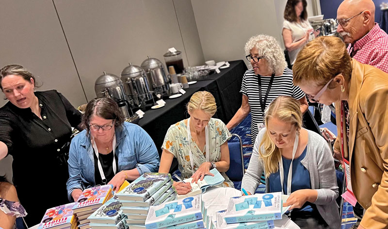 Three women sit at a table autographing piles of books as other people look on.