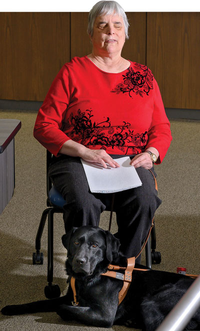 Photograph of Katherine Schneider a woman in a red and black flowered shirt with eye closed touches papers as she reads in braille
